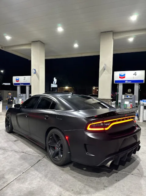 Matte black Dodge Charger with rear spoiler, photographed from behind under a lit fuel station canopy at night