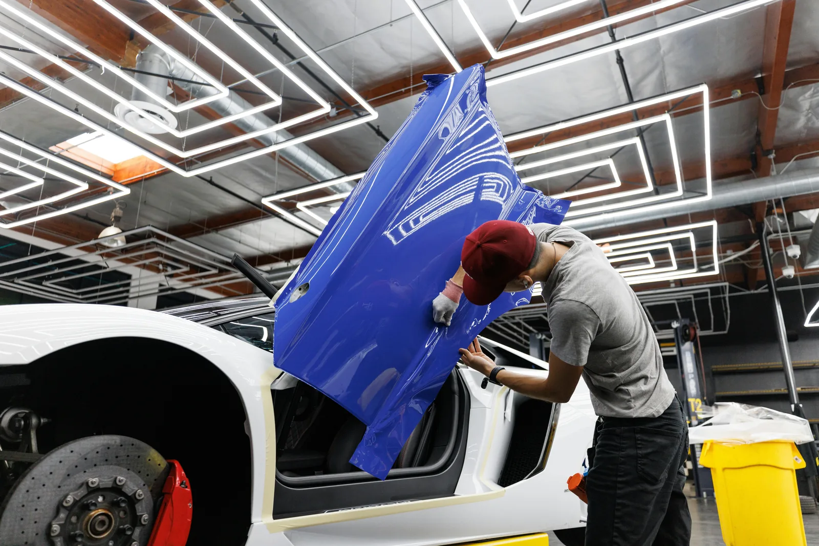 Installer laying a panel of blue gloss vinyl onto the door of a white supercar in a lit wrap bay