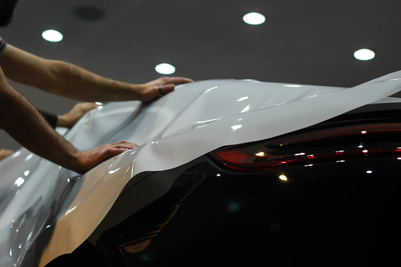 Two hands smoothing a white gloss vinyl panel across the rear quarter of a dark car