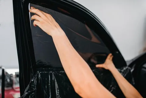 Two hands positioning a sheet of dark window film against a car's rear door glass